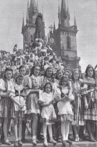 Na podstavci Husova pomníku na Staroměstském náměstí 17. května 1945 (první řada třetí zprava). Spolu s ostatními mávají navrátivším se legionářům, kteří pod nimi defilovali na slavnostní přehlídce. Mezi nimi byl i tatínek paní Tůmové, ale v té chvíli o sobě vzájemně nevěděli. (Zdroj fotografie: Oldřich Smola, „Srdce Prahy v plamenech“, 1946)